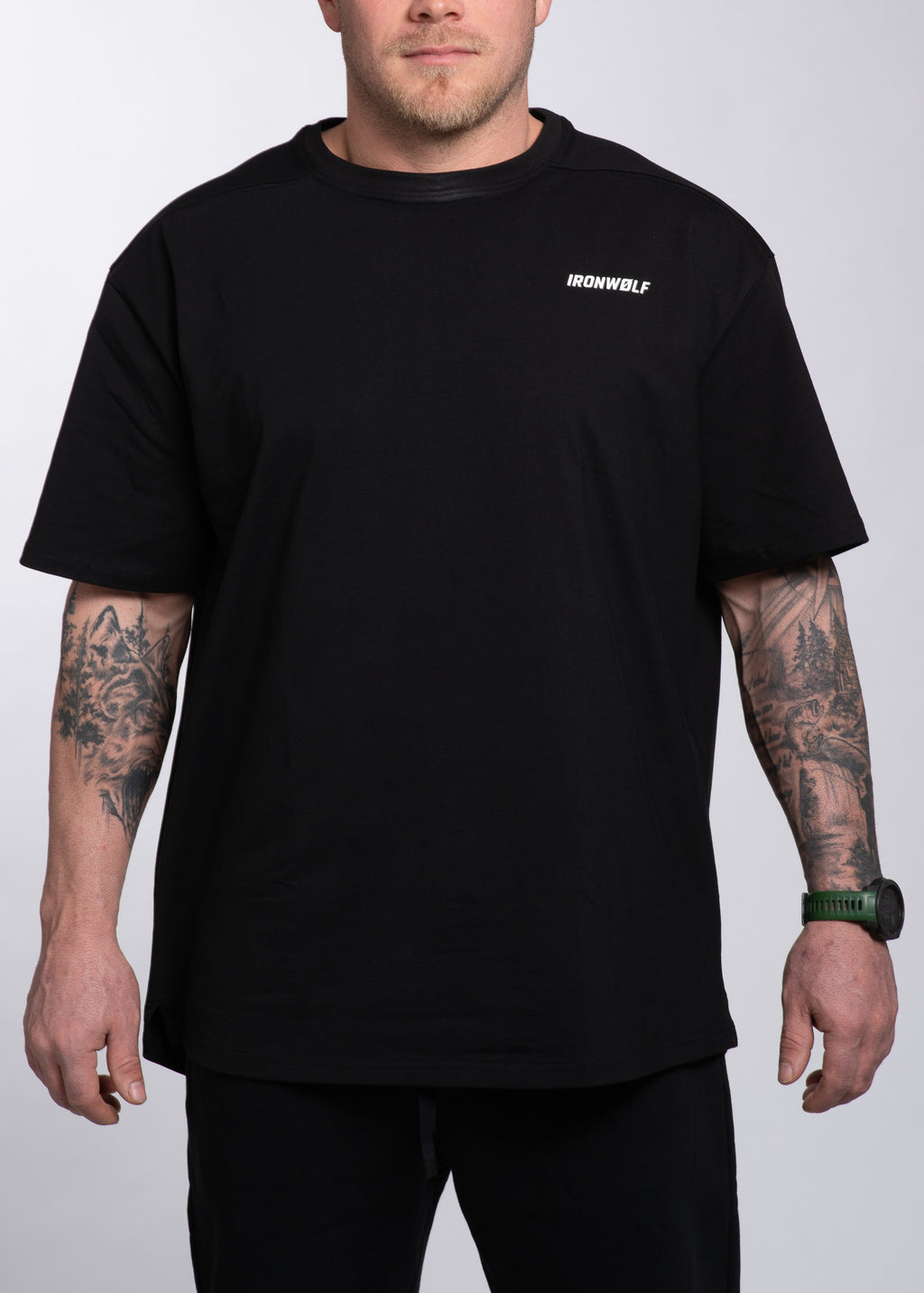Man wearing black IRONWØLF gym t-shirt with chest logo, tattoos, and green watch, styled for training and lifestyle.