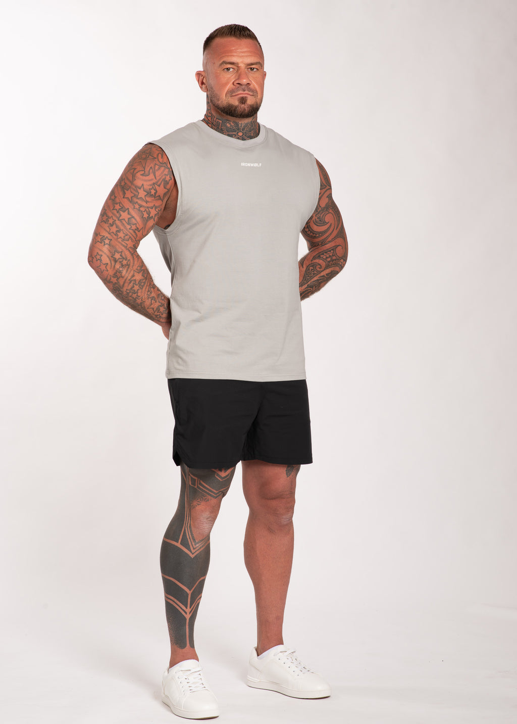 Muscular man in gray sleeveless gym top and black shorts, showcasing tattooed arms and legs for workout wear.