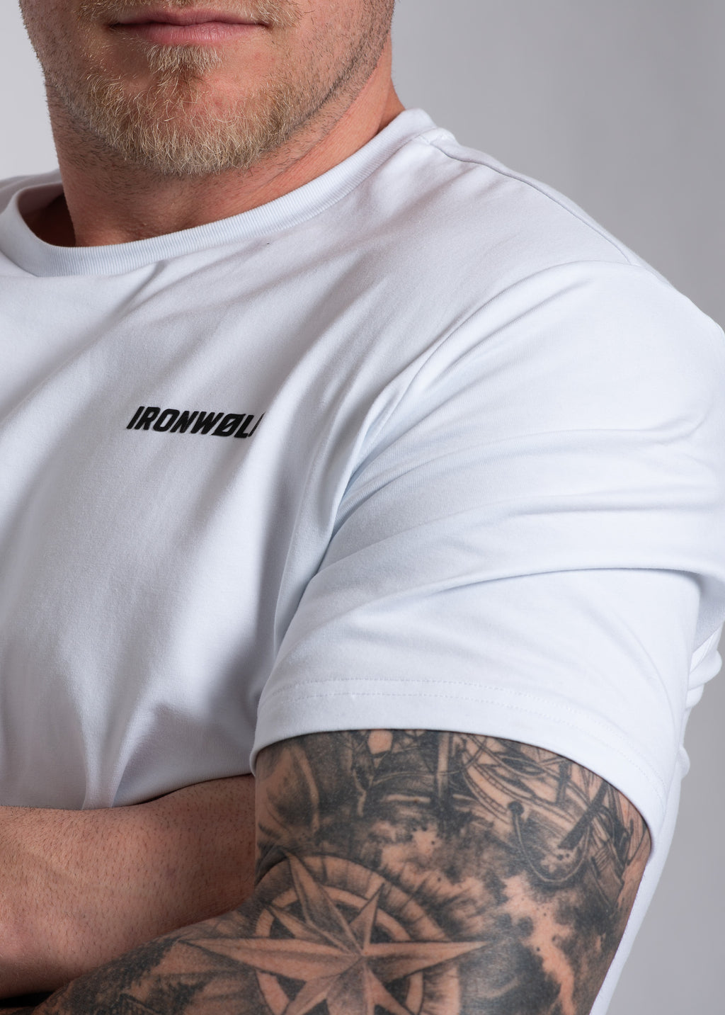 Man wearing white IRONWØLF gym t-shirt with tattooed arm, showcasing athletic activewear for training and lifestyle.