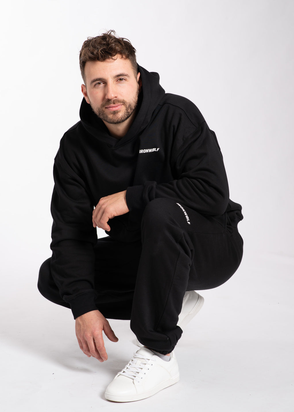 Man in black IRONWØLF hoodie and joggers, crouching on white background, wearing white sneakers