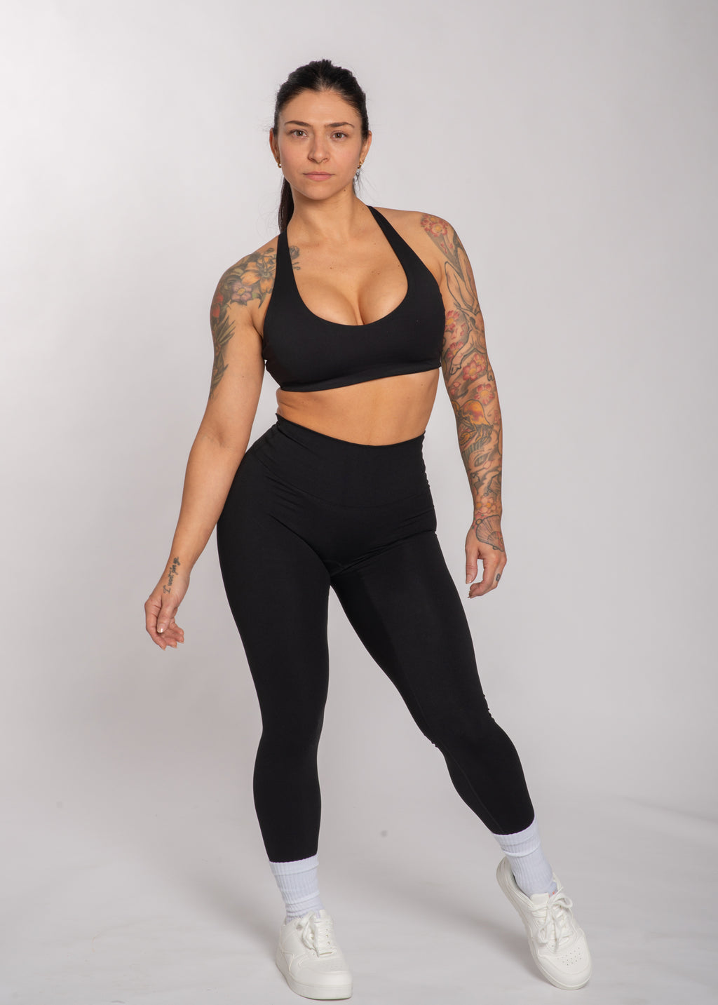 Woman in black sports bra and high-waisted leggings, tattooed arms, white sneakers, gym wear