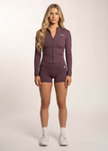 Woman in purple long sleeve zip-up gym top and matching shorts, white sneakers, studio background