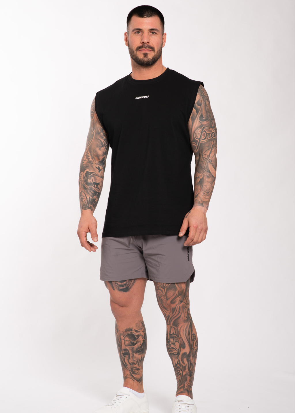 Man in black sleeveless gym shirt and gray shorts, showcasing tattooed arms and legs for workout wear.