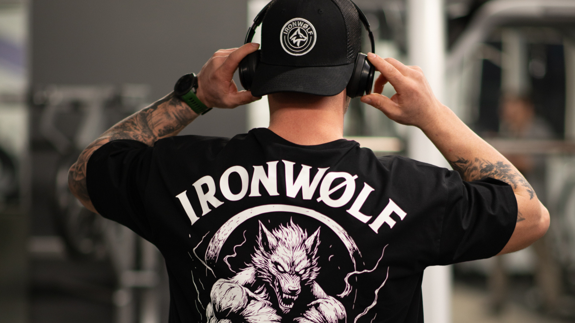 Man in gym wearing IRONWØLF graphic tee and cap, adjusting headphones for workout