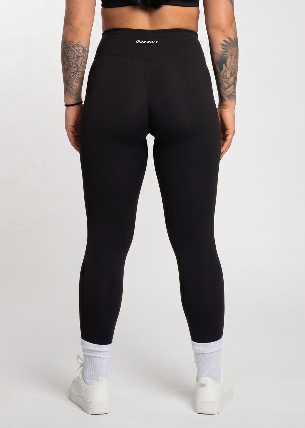 Woman wearing black high-waisted gym leggings with IRONWØLF waistband, white socks, and sneakers