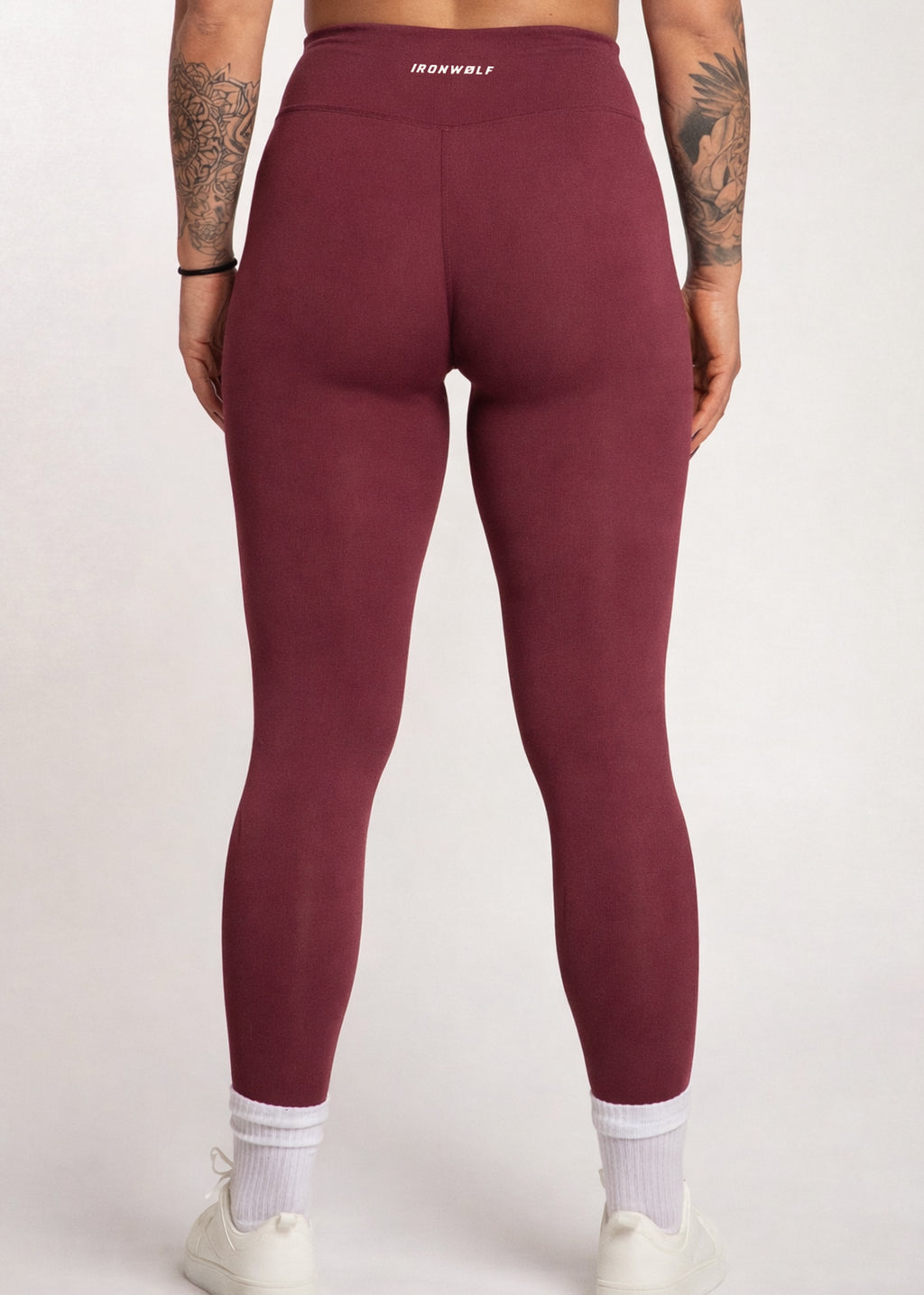 Woman wearing burgundy high-waisted gym leggings with IRONWØLF logo, white socks, and sneakers